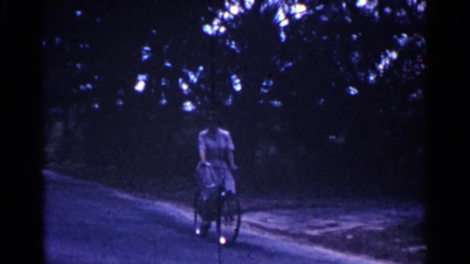 The woman continues riding her bicycle along a curving Nassau road, partly obscured by deep shade and soft 8mm focus.