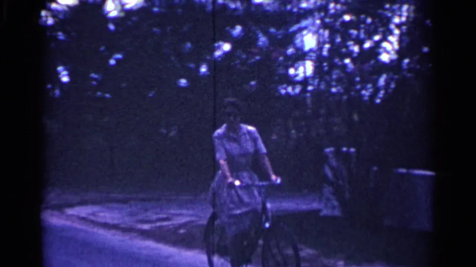 A woman in a white dress rides a bicycle near a low wall and shaded trees in 1963 Nassau, Bahamas.