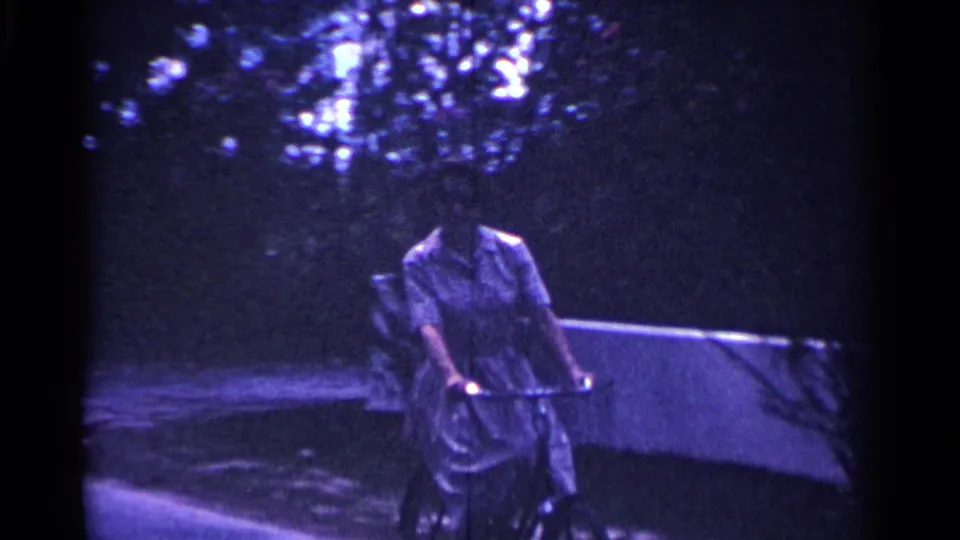 In 1963 Nassau, a woman in a light dress rides a bicycle with a basket along a shaded sidewalk.