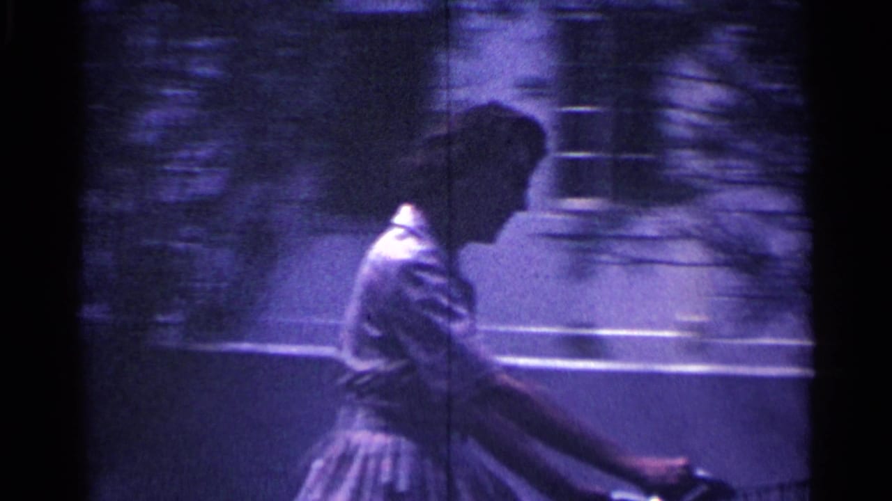 Side view of the woman riding her bicycle along a Nassau sidewalk, with a low wall and trees behind her.