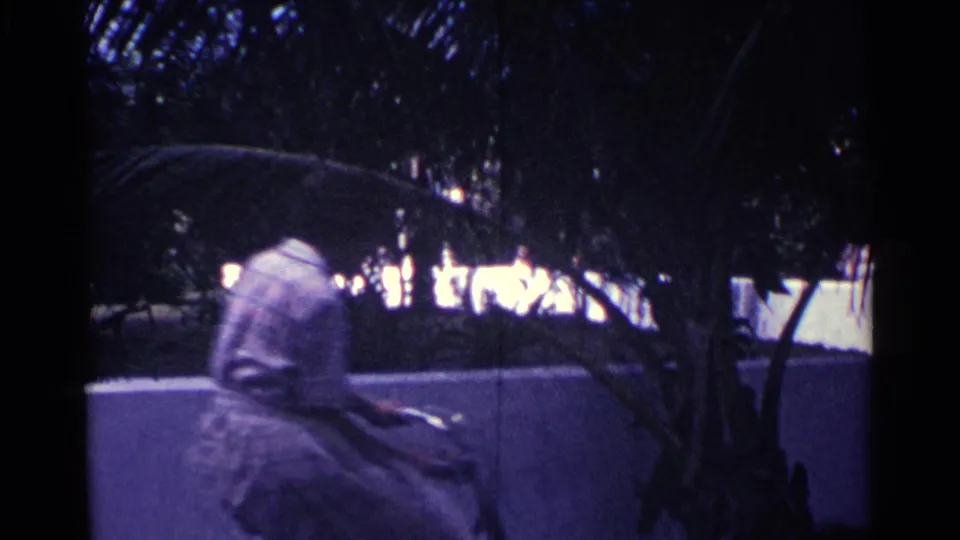The woman cycles away from the camera under tree cover, continuing along the sidewalk in Nassau.