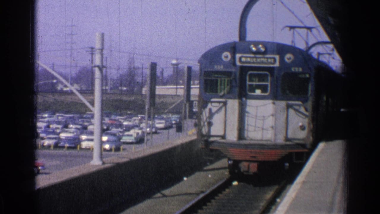 1965 Ohio home-movie frame of a blue-and-white passenger train approaching a station platform near a parking lot of period cars.
