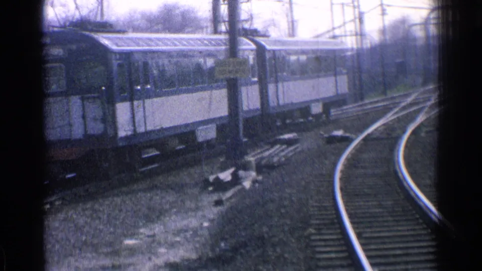 Blue-and-white passenger cars move along a curved electrified track in Ohio, seen through soft 8mm film grain.