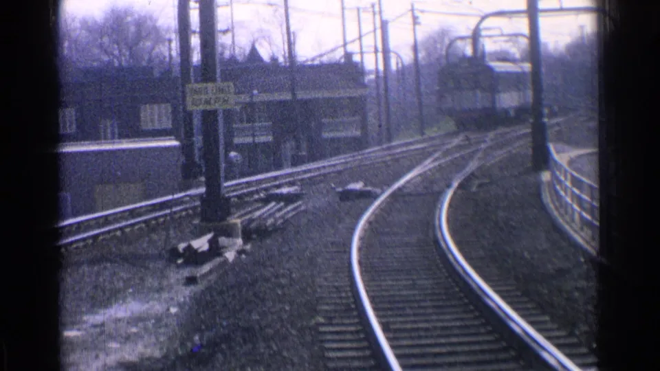 1965 Ohio rail scene with curved tracks, overhead electrical supports, platform railings, and station buildings in a soft 8mm frame.