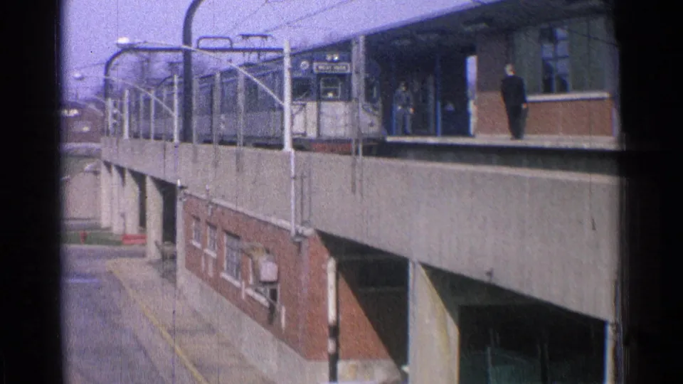 1965 Ohio 8mm frame showing an elevated station platform, metal railings, brick support walls, signage, and a few distant people.