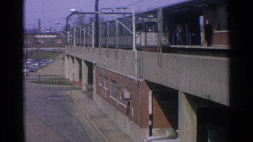 1965 Ohio station view showing platform railings, a brick-and-concrete structure, posted signs, and distant people near the building.