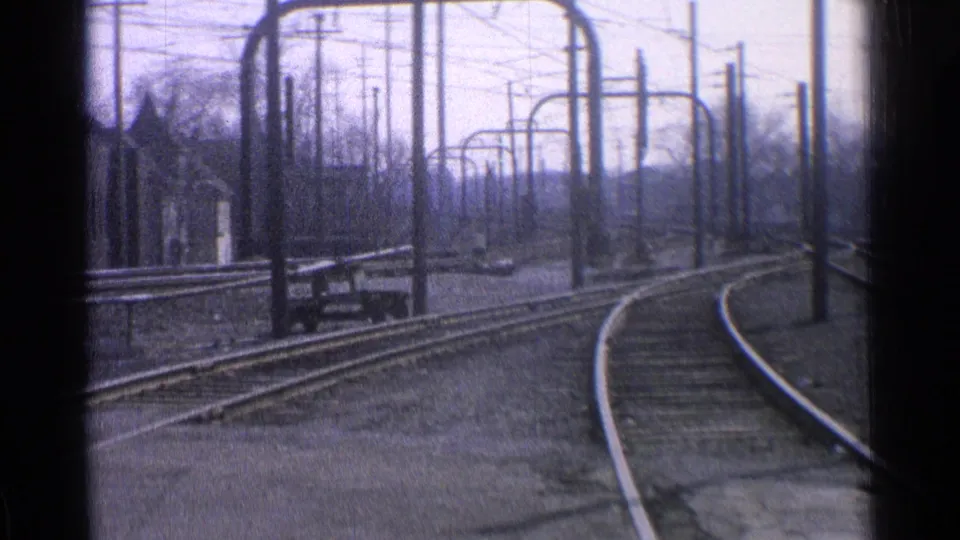 1965 Ohio home-movie frame of curved rail tracks, overhead catenary supports, utility poles, and bare trees in a quiet train yard.
