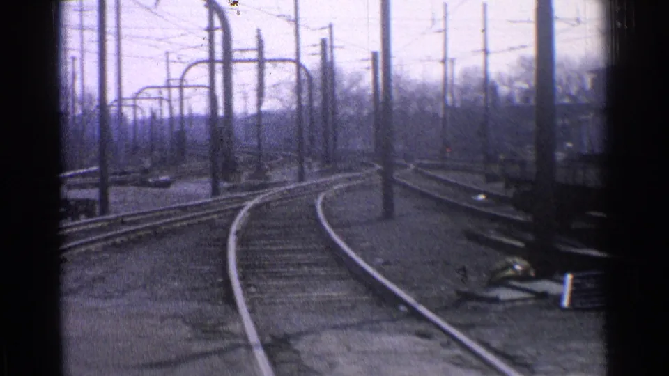 1965 Ohio 8mm frame showing curved tracks, overhead line poles, and the edge of a trolley or rail car at the right side of the yard.