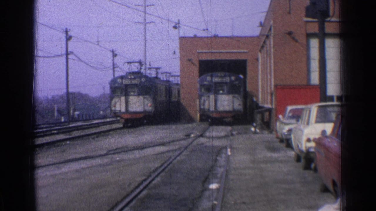 Two trolley cars near a brick maintenance garage with tracks, overhead wires, and parked cars.