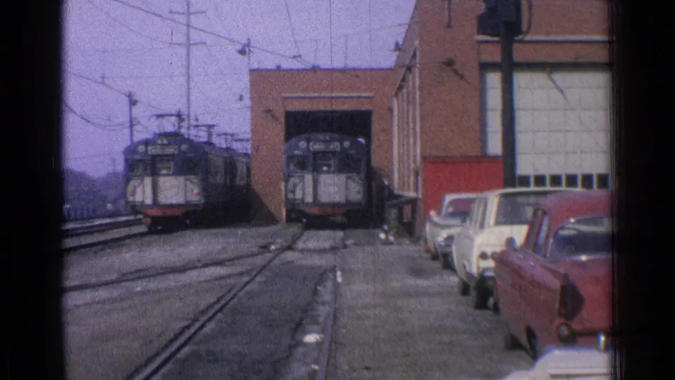 1965 Ohio street-level view of trolley cars at an industrial maintenance facility with adjacent parked cars and overhead electric lines.