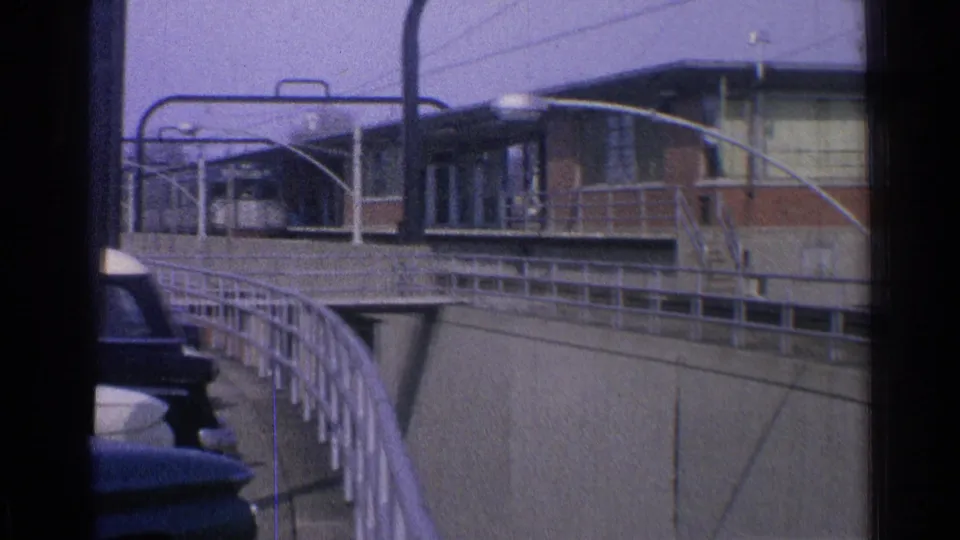 1965 Ohio 8mm frame of a trolley beside a maintenance structure, seen across a curved railing with streetlights and parked cars at the edge.