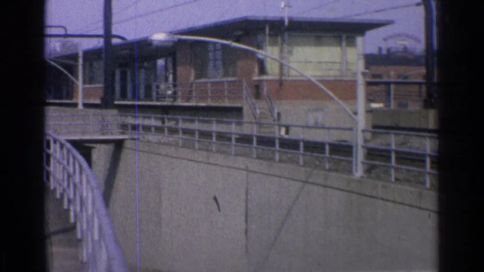 1965 Ohio view of a trolley at a maintenance yard, with a raised walkway railing, streetlights, overhead wires, and industrial buildings.