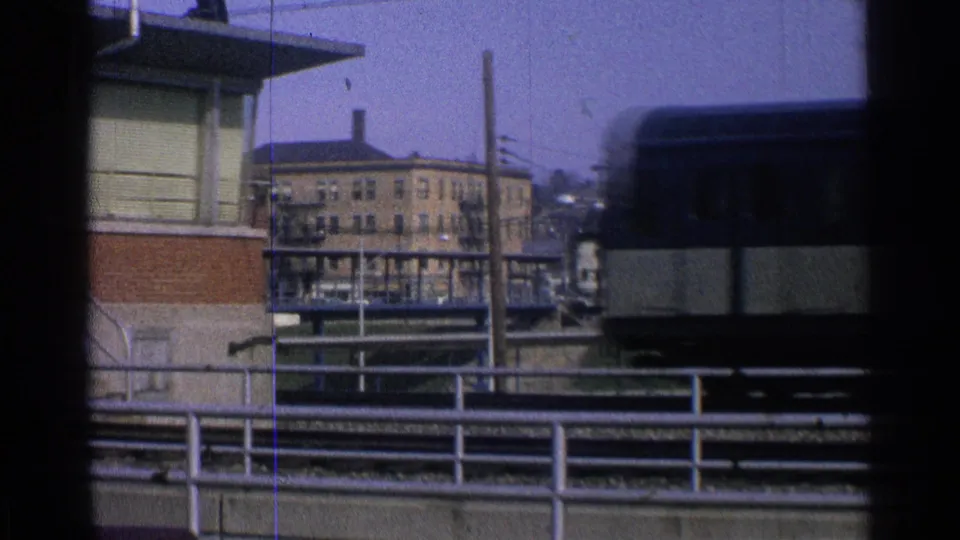 Blue and cream passenger train entering the frame on an elevated track in an Ohio urban setting, with railings and buildings behind it.