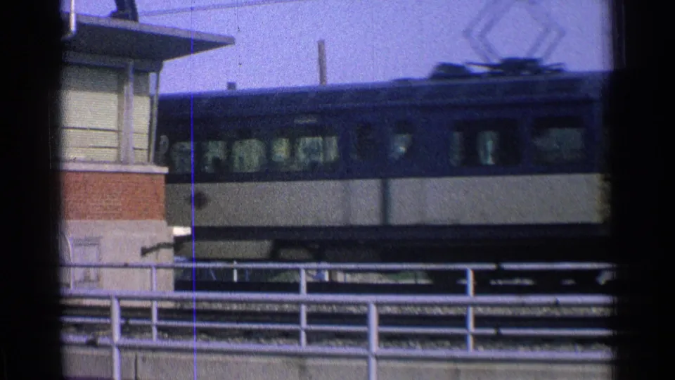1965 Ohio home-movie frame of a blue and cream passenger railcar moving across an elevated urban track with railings below.