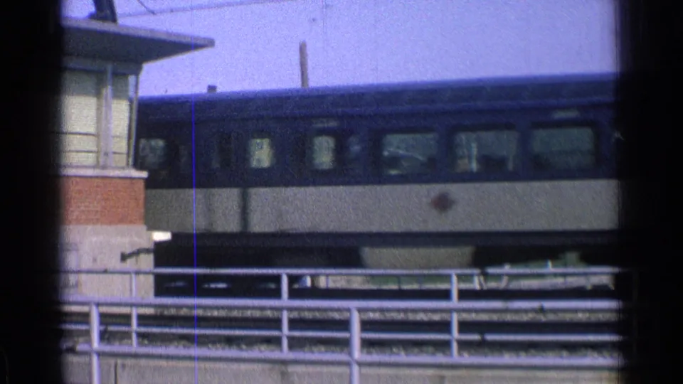 Blue and cream passenger train continuing across an elevated Ohio urban rail line, framed by dark window edges and 8mm film texture.