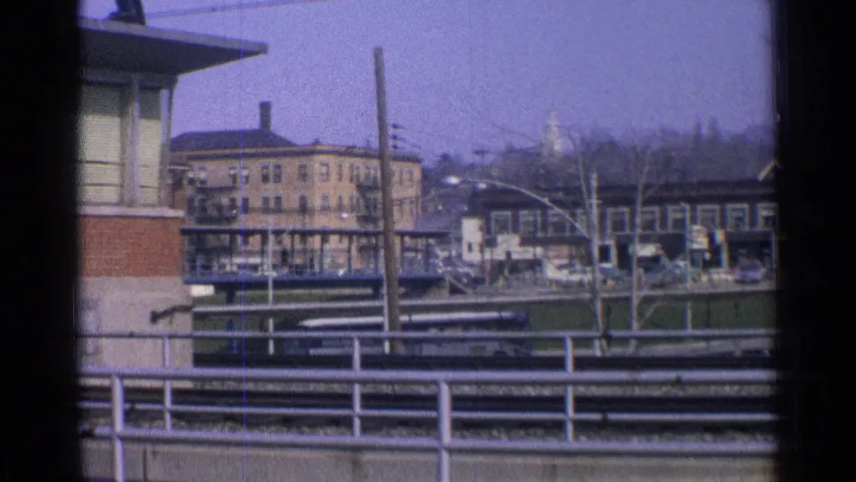 1965 Ohio 8mm frame with elevated rail tracks, station-area buildings, railings, utility poles, and leafless trees under a hazy sky.