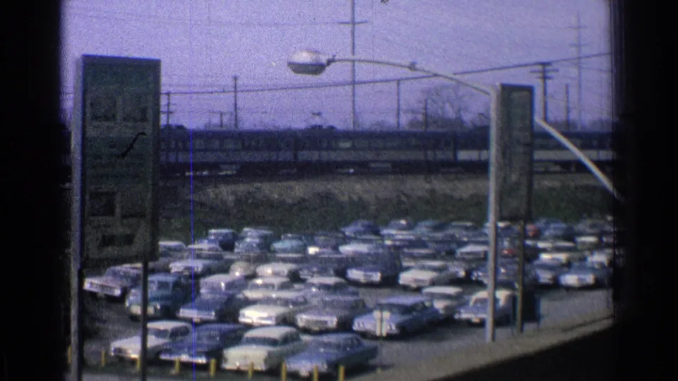 1965 Ohio 8mm frame showing a busy parking lot of mid-century cars below elevated tracks, with a train visible beyond streetlights and poles.