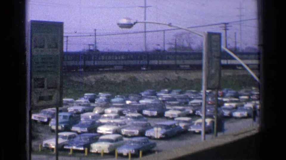1965 Ohio 8mm frame of a crowded lot with 1960s sedans and station wagons, elevated railway, streetlights, poles, and bare trees.