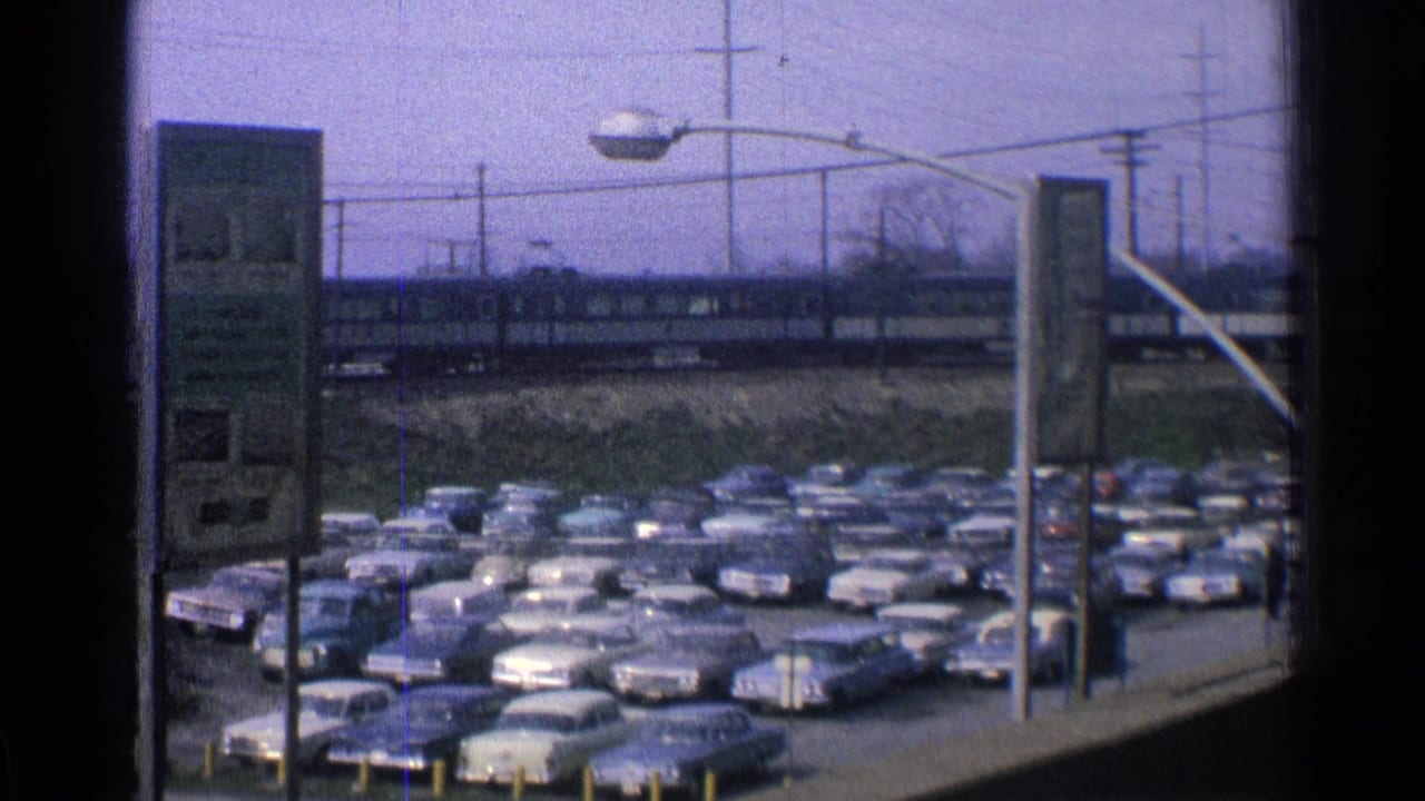 Crowded station parking lot with classic cars and a passenger train passing behind it.
