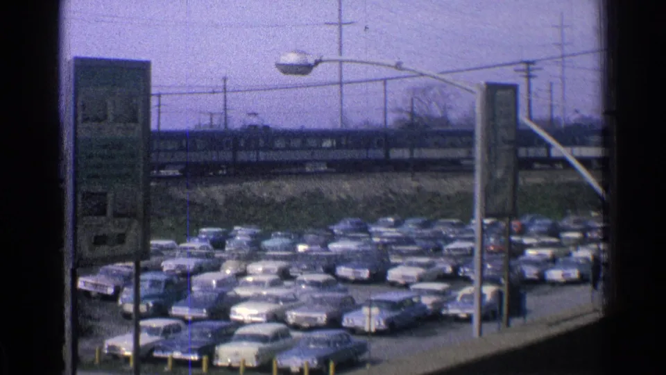 1965 Ohio home-movie view of classic cars in a station parking lot with a passenger train on elevated tracks in the background.
