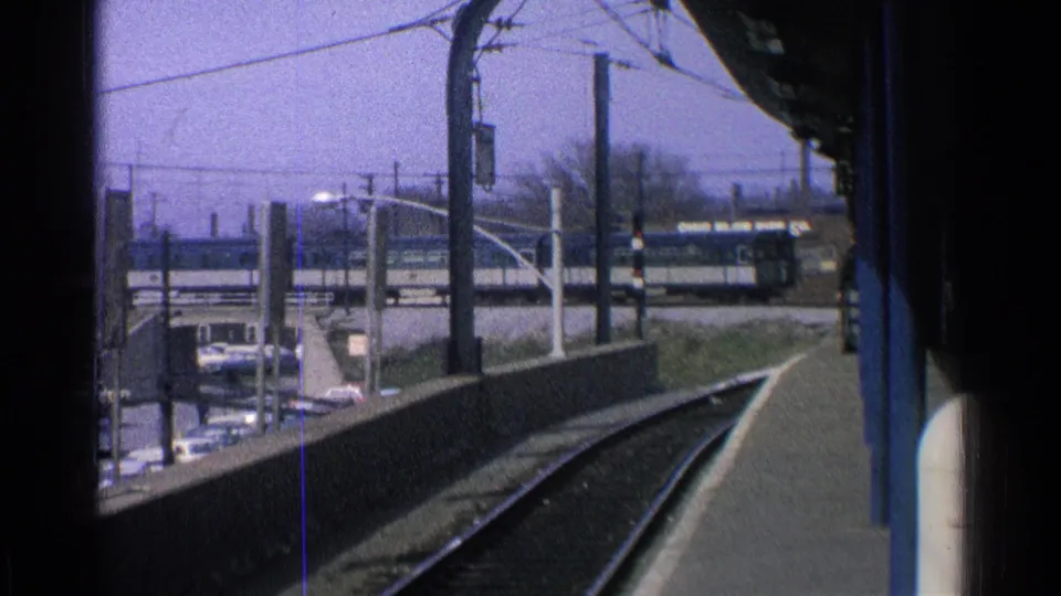 1965 Ohio home-movie frame from a station platform, with rails curving ahead and a passenger train beyond the parked cars.