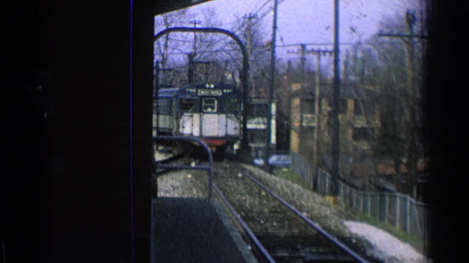 1965 Ohio station approach showing a passenger train coming toward the camera along tracks bordered by fencing and winter-bare trees.