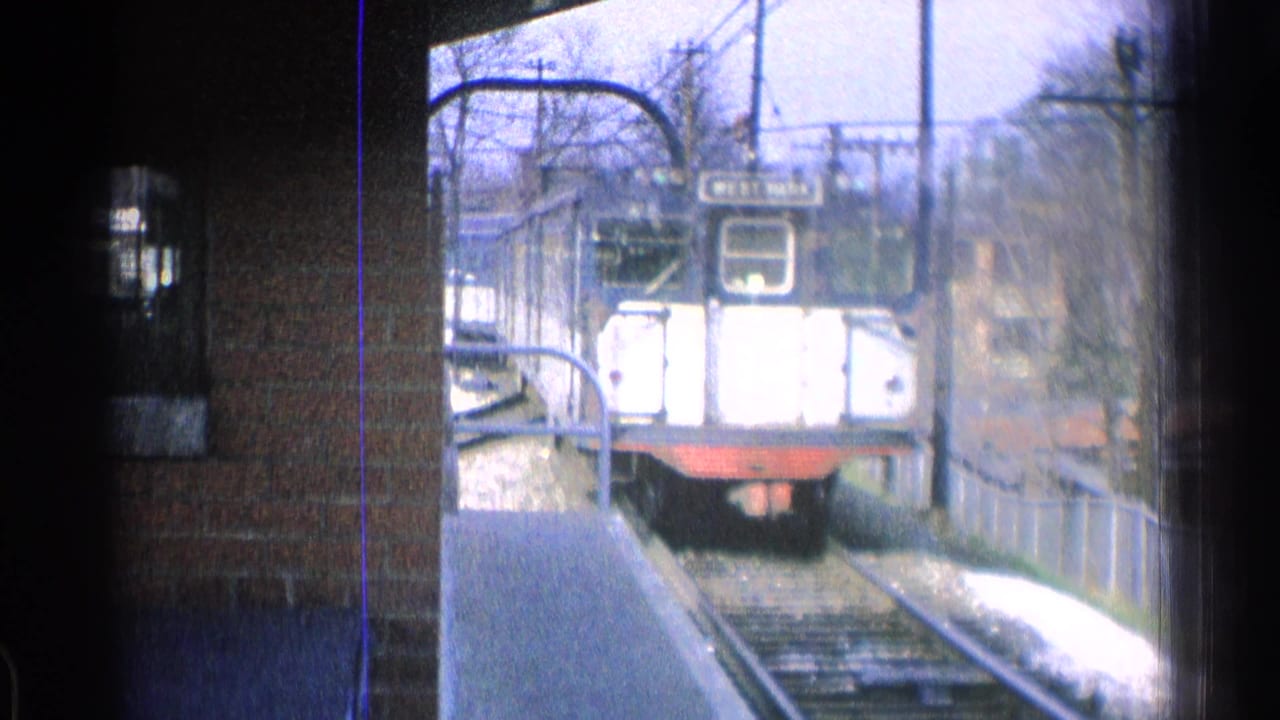1965 Ohio 8mm frame of a dark blue and white train approaching along tracks beside a brick wall and platform railing.