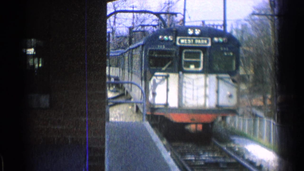 The train nears the station platform in grainy 1965 Ohio home-movie footage, with bare trees and overhead lines behind it.
