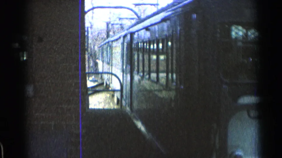 The train continues past the platform in a soft, grainy 1965 Ohio home-movie frame with railings, brickwork and overhead wires visible.