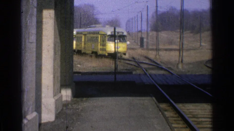 1965 Ohio 8mm frame of yellow rail cars near a station platform, with converging tracks, overhead poles, and bare rural trees.