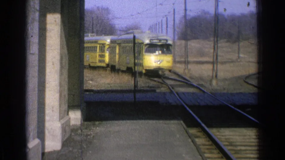 1965 Ohio frame showing the front and side of a yellow train near a platform, with tracks, poles, and bare trees in the background.