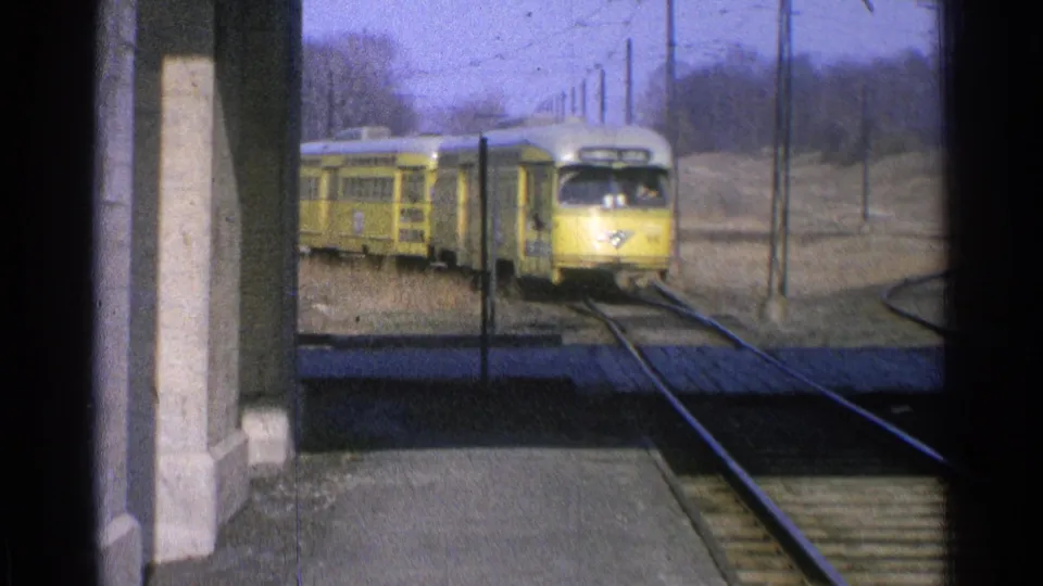 1965 Ohio 8mm frame of a yellow electric train approaching a station platform under overhead wires.