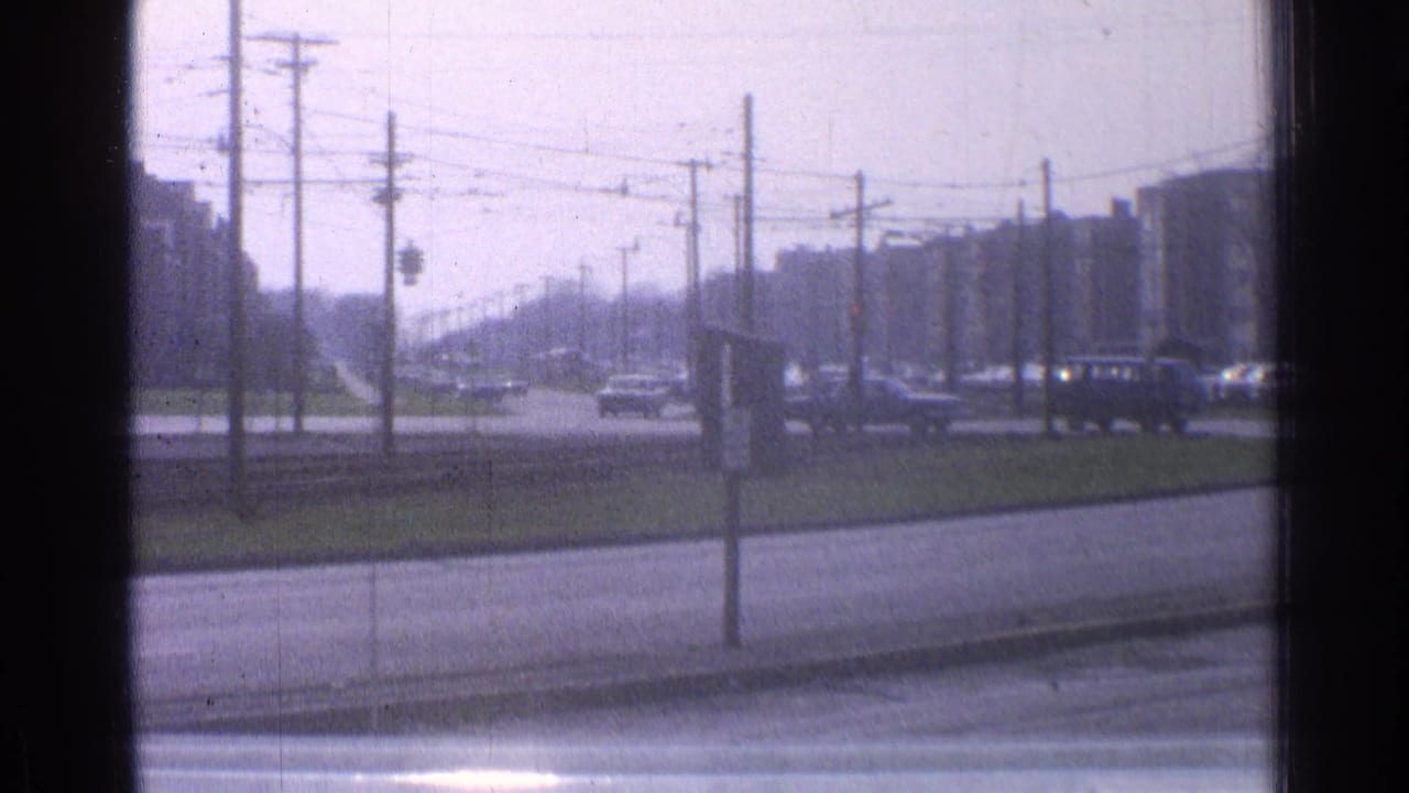 1965 Ohio street view from a moving vehicle, with cars crossing a broad road beneath overhead wires near apartment blocks.