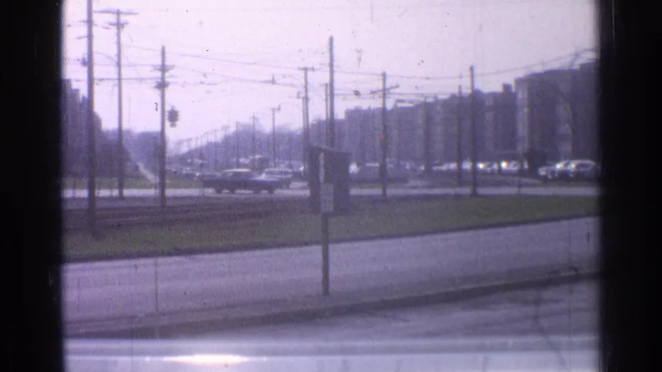 1965 Ohio home-movie frame showing cars on a wide urban road, seen through a soft, dusty vehicle window.