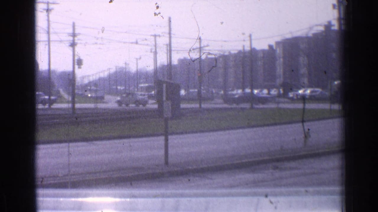 1965 Ohio street view with light traffic, overhead wires, a grassy median, and distant mid-rise buildings seen through a window frame.