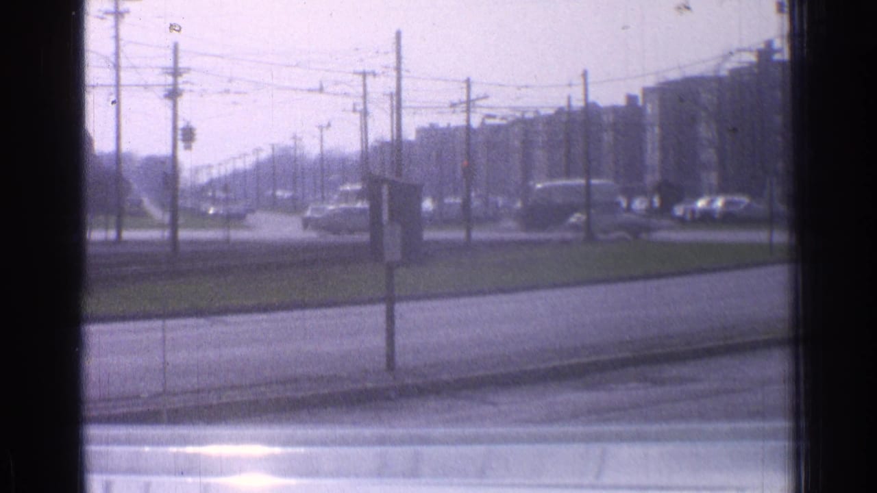 1965 Ohio downtown traffic scene with classic cars moving beneath overhead wires near a grassy median and mid-rise buildings.