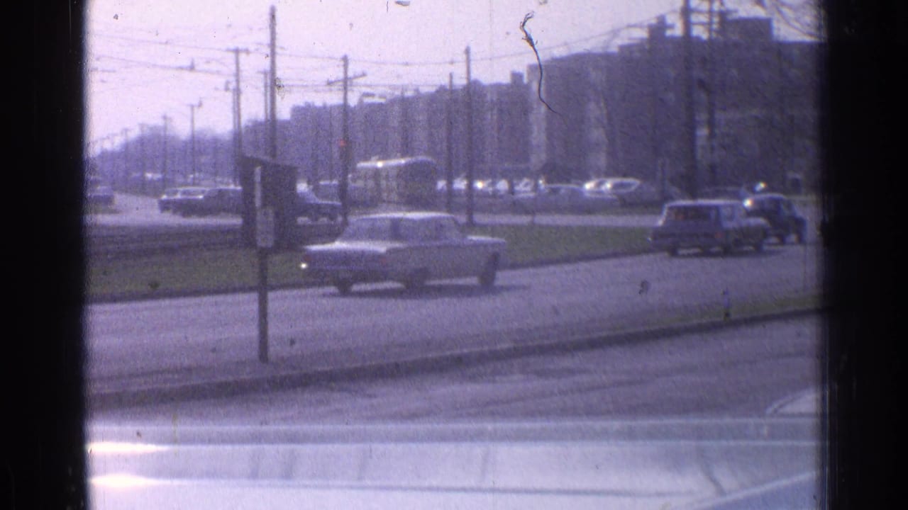 Daytime 1965 Ohio street traffic with mid-century cars crossing near utility poles, overhead wires, and distant urban buildings.