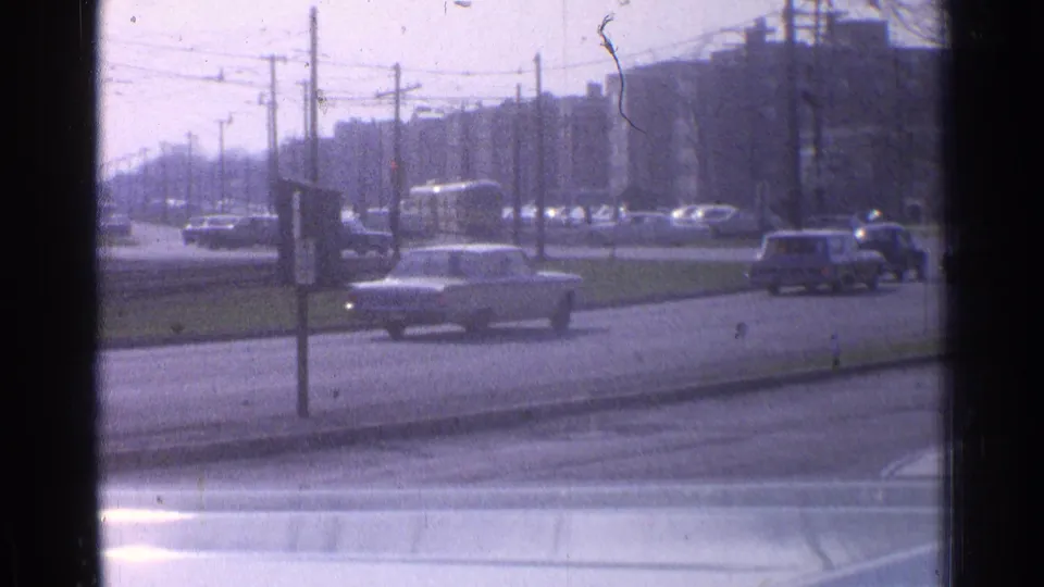 Daytime 1965 Ohio street traffic with mid-century cars crossing near utility poles, overhead wires, and distant urban buildings.