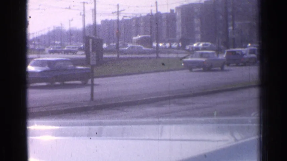 1965 Ohio city traffic viewed through glass, showing several cars, roadside posts, overhead wires, and hazy mid-rise buildings.