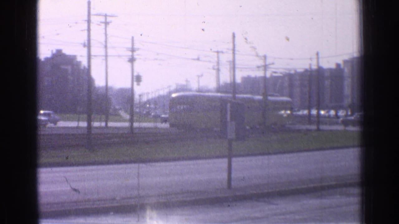 1965 Ohio home-movie frame showing a yellow train beyond a roadside, with utility poles, wires, traffic signals, and distant buildings.