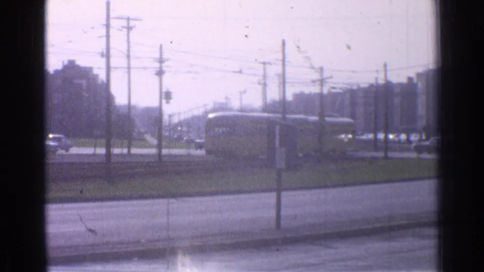 1965 Ohio home-movie frame showing a yellow train beyond a roadside, with utility poles, wires, traffic signals, and distant buildings.