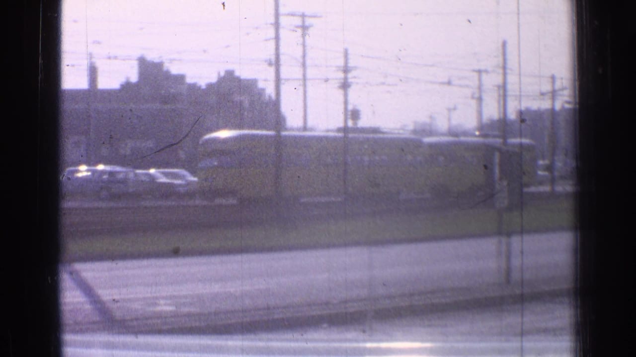1965 Ohio frame with a yellow railcar moving past parked cars, utility poles, overhead wires, and low town buildings.