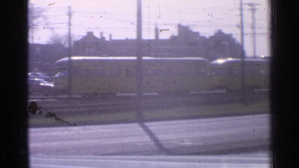 1965 Ohio home-movie frame showing a long yellow train beside a road, with utility poles, tracks, and town buildings in the background.