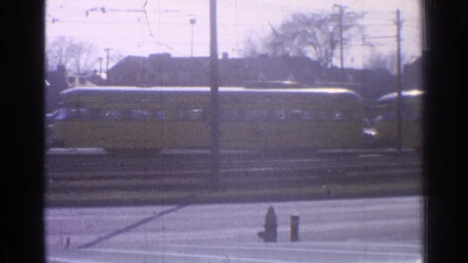 1965 Ohio frame of a yellow train moving behind road lanes and tracks, with utility poles, bare trees, distant buildings, and a hydrant visible.