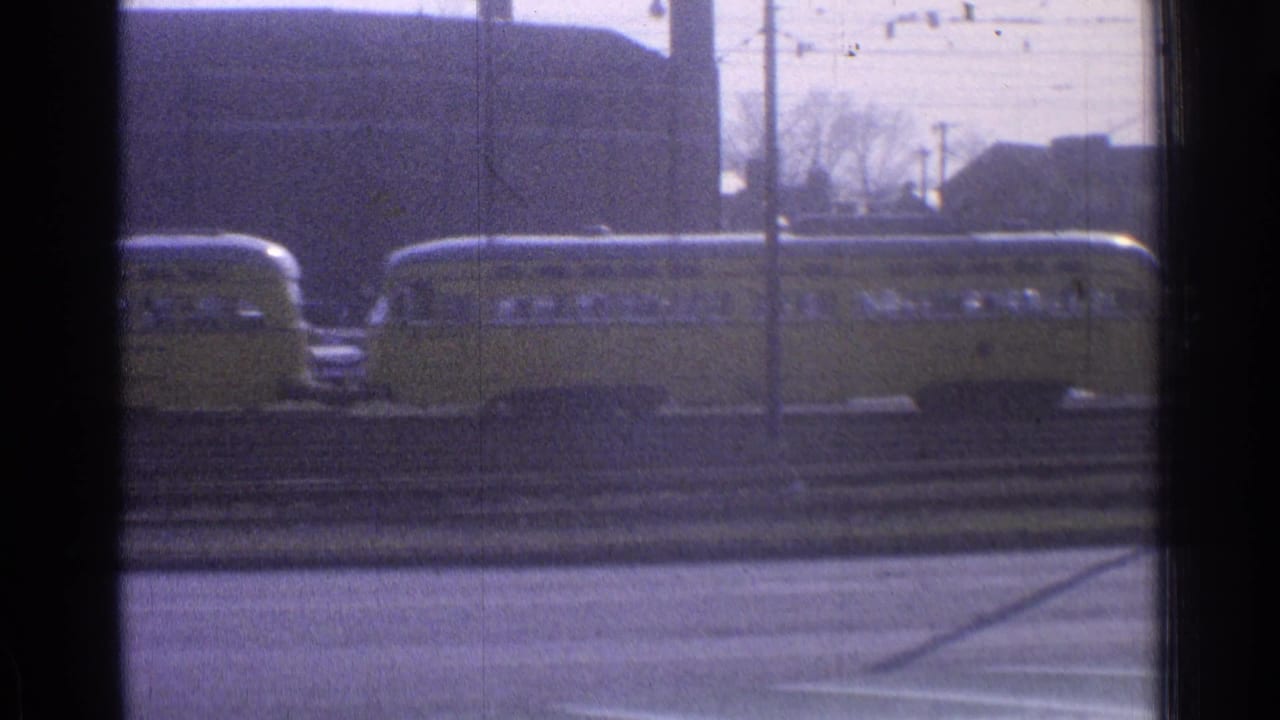 Yellow passenger railcars pass in front of an industrial building and utility lines.