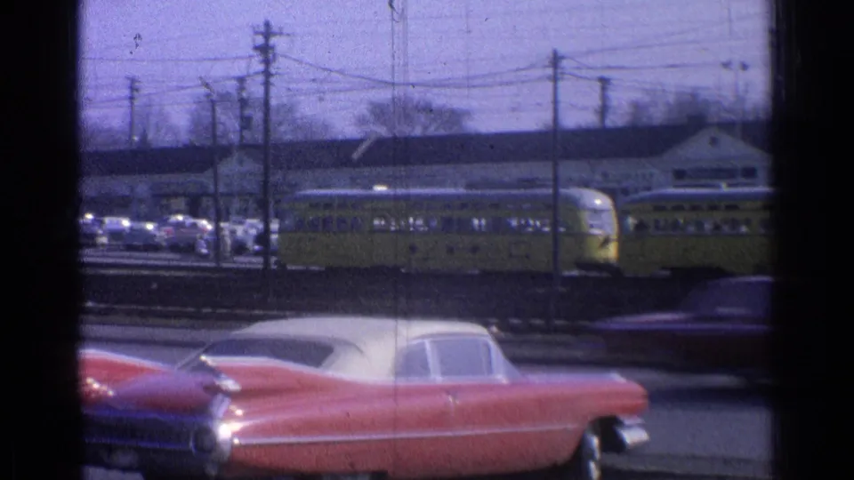 1965 Ohio 8mm frame with yellow passenger railcars, roadside traffic, utility poles, and a red mid-century car in the foreground.