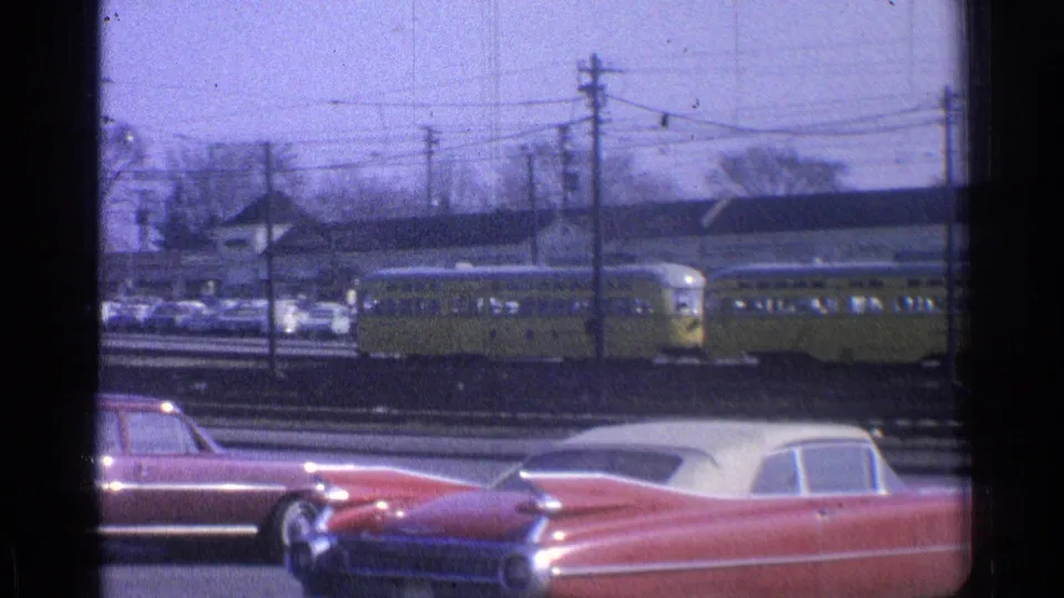 1965 Ohio 8mm frame showing a yellow passenger train behind classic cars, tracks, utility poles, and low industrial buildings.