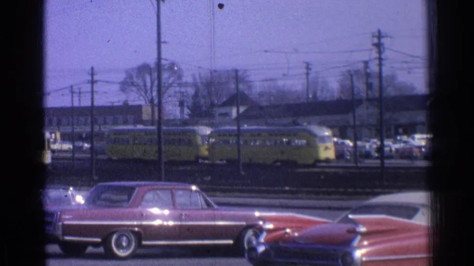 1965 Ohio 8mm frame showing two yellow trolleys, roadside classic cars, utility poles, and low buildings in a suburban setting.