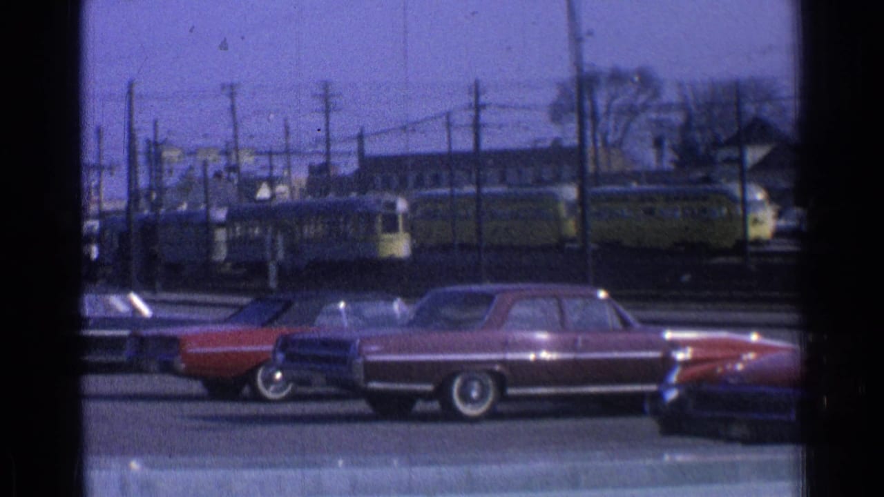 1965 Ohio home-movie frame showing parked sedans with a yellow trolley or light rail vehicle beyond the lot.