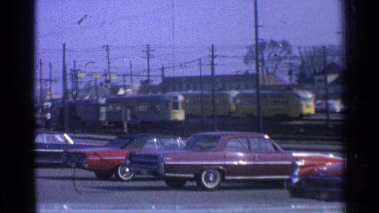 1965 Ohio street view through a dark window frame, with sedans parked before a yellow rail vehicle and overhead wires.